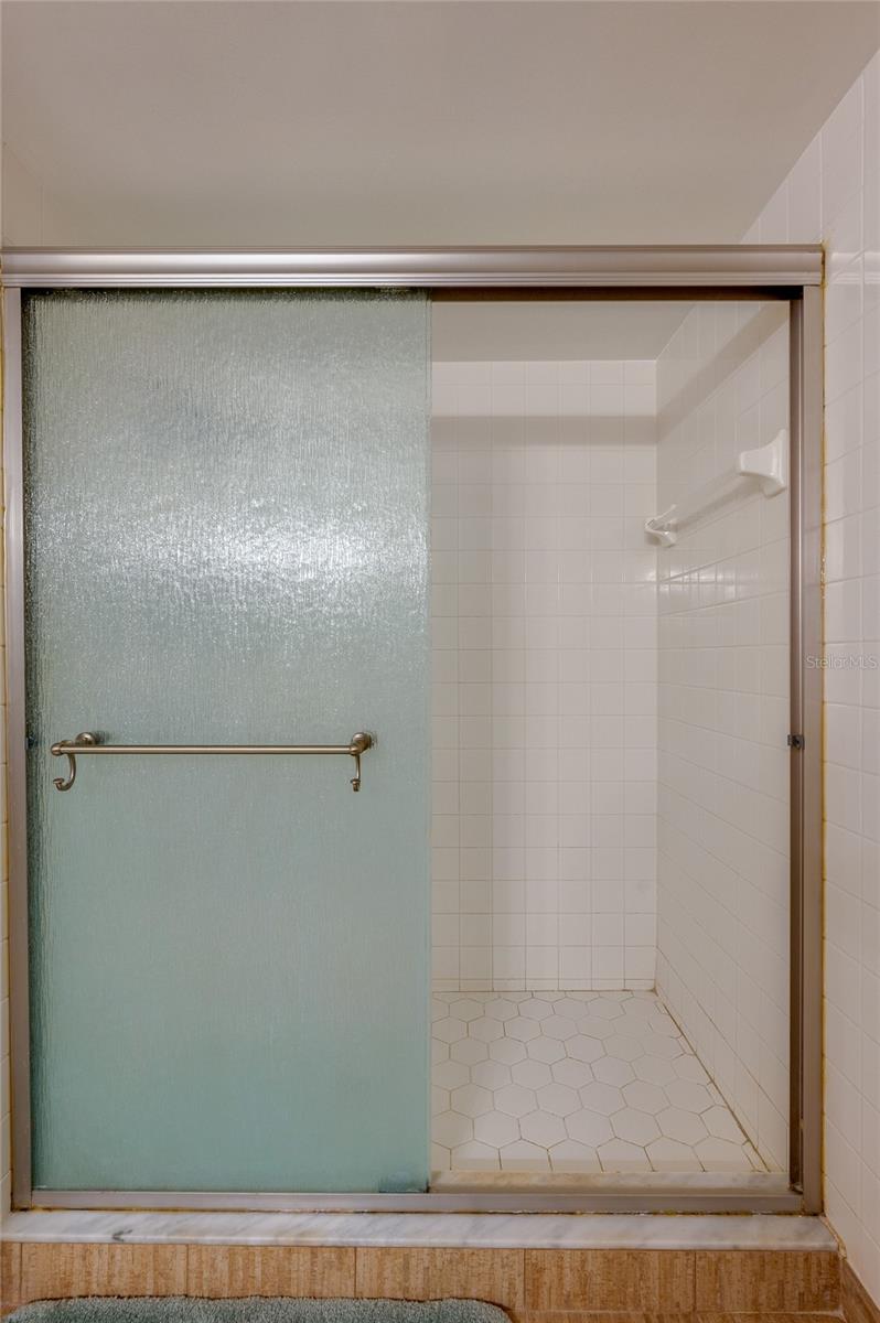 Close-up view of the primary suite’s shower showcases a hexagon tile floor and full tile surround behind frosted sliding doors. - 6727 TURTLEMOUND RD #315, NEW SMYRNA BEACH