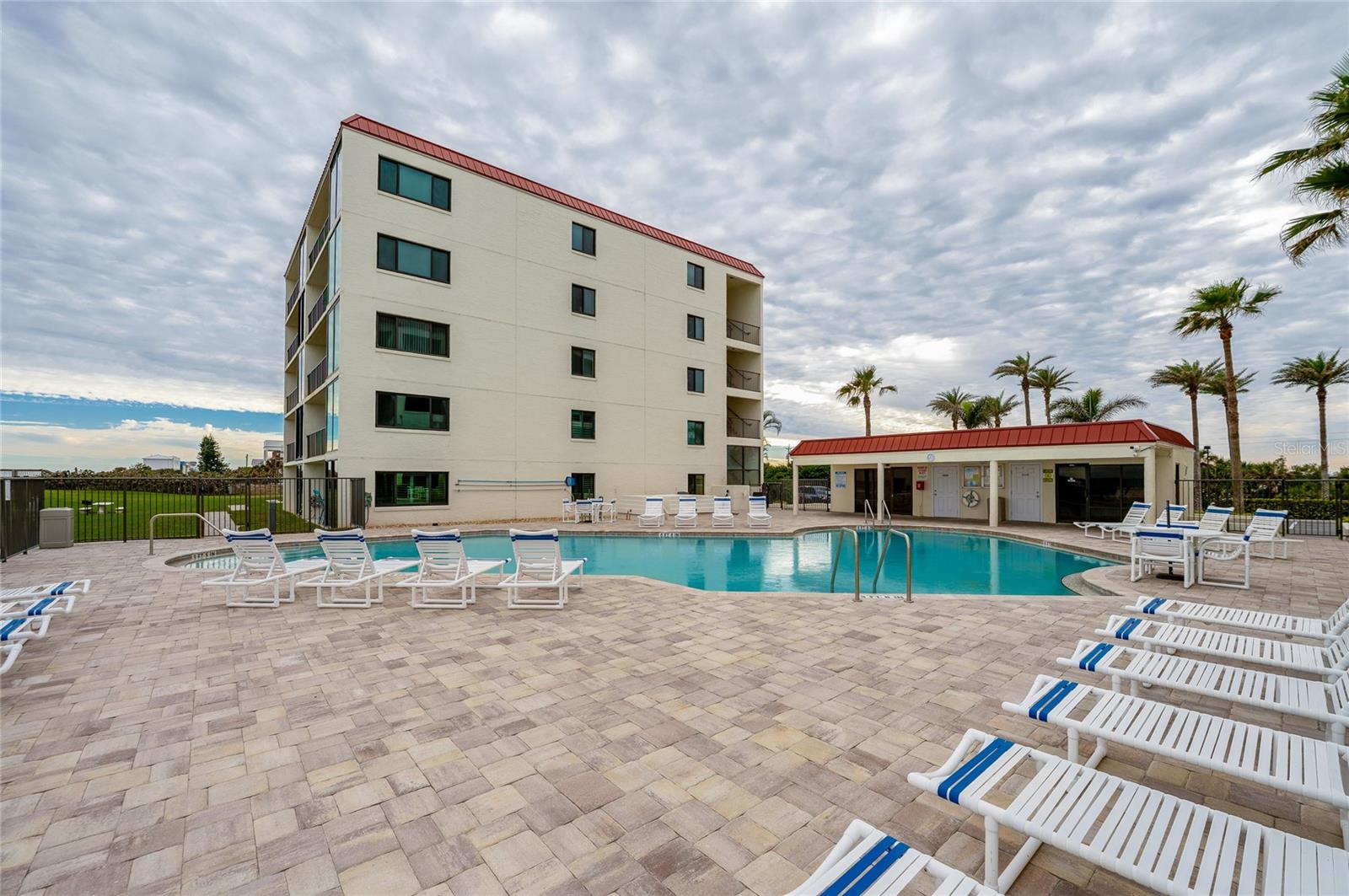 The resort-style pool includes a spacious deck, poolside lounge chairs, and a nearby clubhouse building, all set against a backdrop of palm trees and ocean views. - 6727 TURTLEMOUND RD #315, NEW SMYRNA BEACH