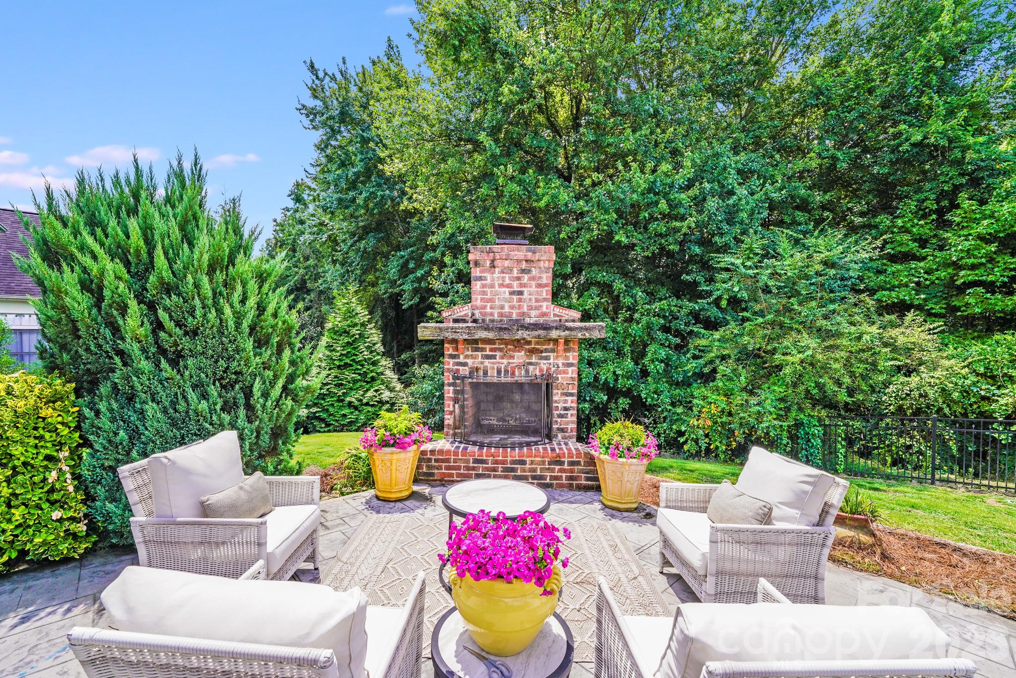 This inviting outdoor living area features a classic brick fireplace surrounded by lush landscaping—perfect for cozy gatherings or peaceful evenings under the stars - 2702 Arsdale Road, Waxhaw