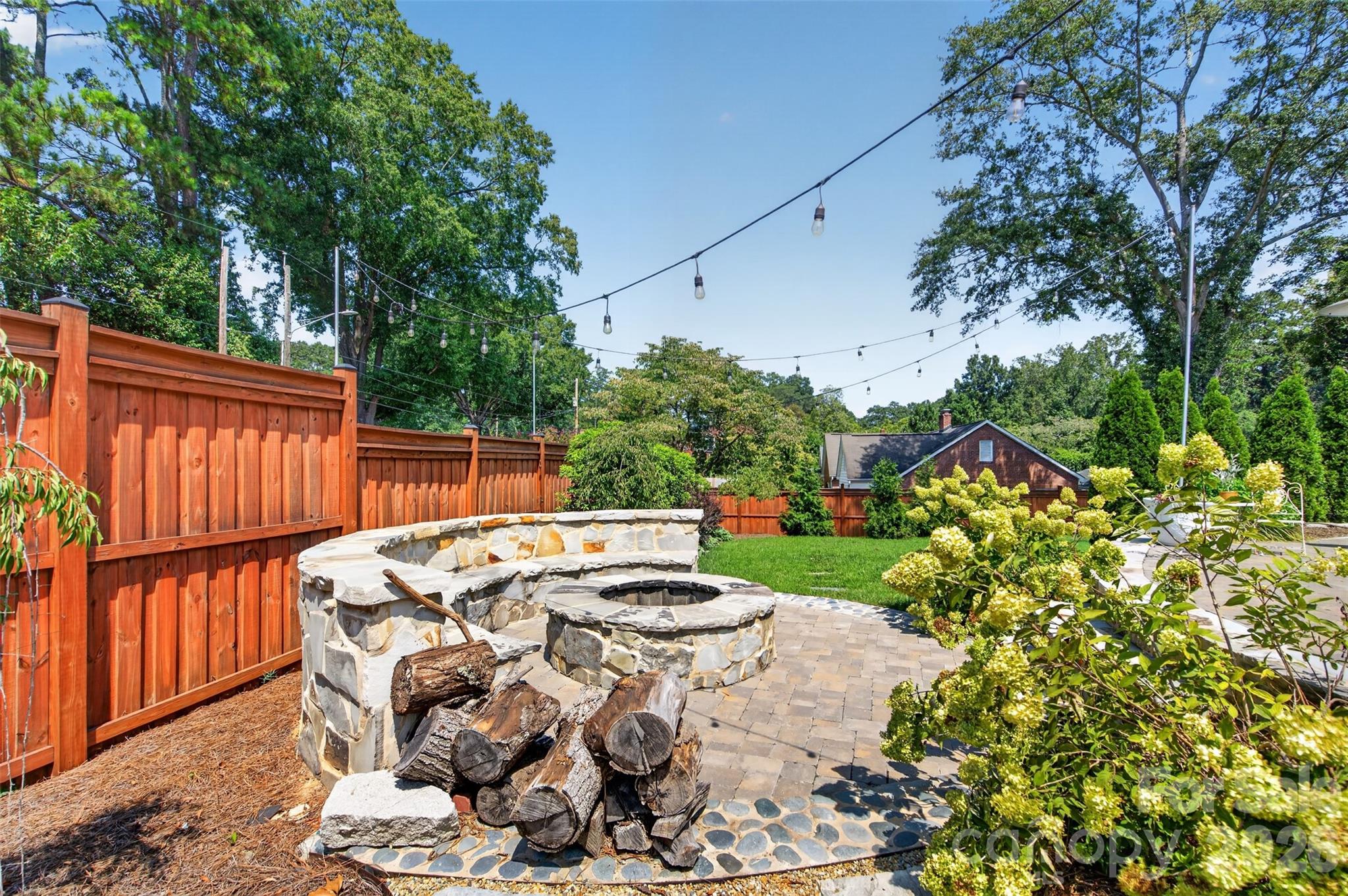 A dedicated fire pit area with stone seating, surrounding landscaping, and overhead string lights creates another inviting spot to gather outdoors. - 638 Carolina Avenue, Gastonia
