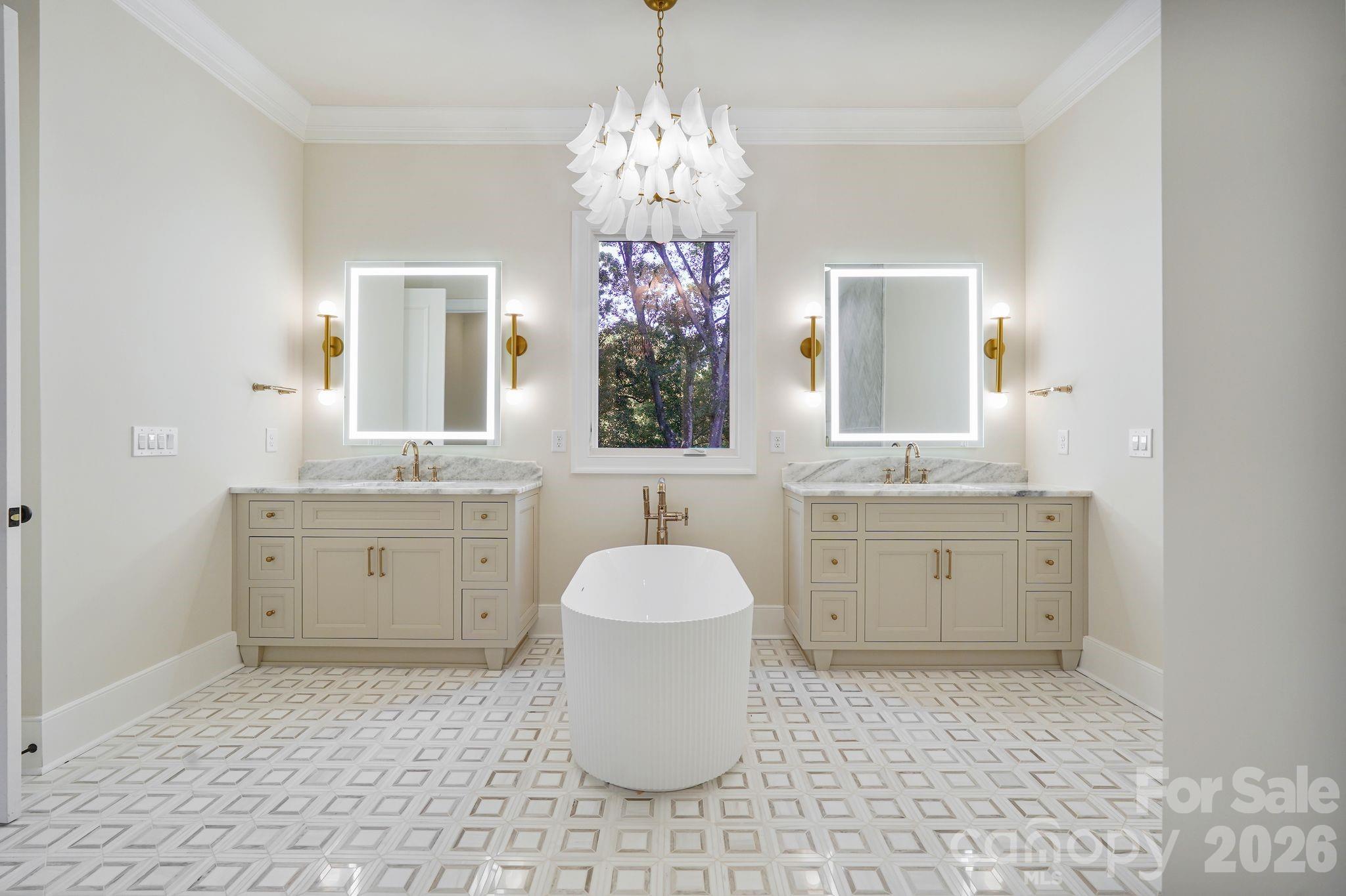 Spa-inspired primary bath featuring separate his-and-hers vanities, a sculptural freestanding tub with statement lighting, luxury illuminated mirrors, and a large window that fills the space with natural light. - 21216 Rio Oro Drive, Cornelius