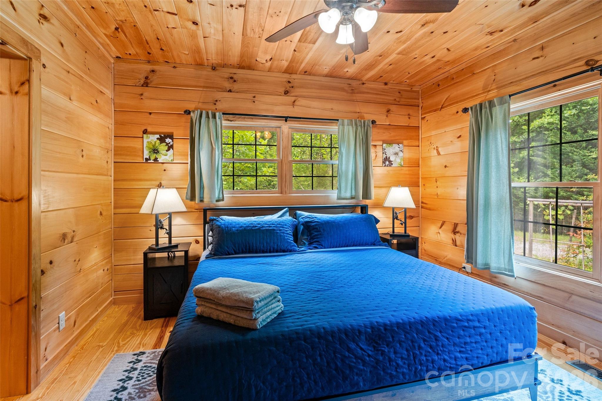 Retreat to this tranquil primary bedroom, where floor-to-ceiling wood paneling creates a warm, cabin-cozy ambiance. Multiple windows let in plenty of natural light and frame beautiful, leafy views of the surrounding forest to wake up to each morning. - 213 Big Valley Road, Pisgah Forest