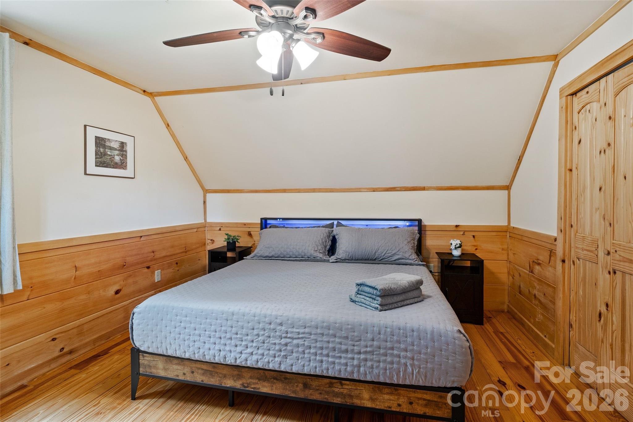 Spacious upper-level bedroom boasting charming rustic details, including natural pine floors, wood wainscoting with matching trim, an angled ceiling line, integrated lighting, a ceiling fan, and double panel wood closet doors. - 213 Big Valley Road, Pisgah Forest