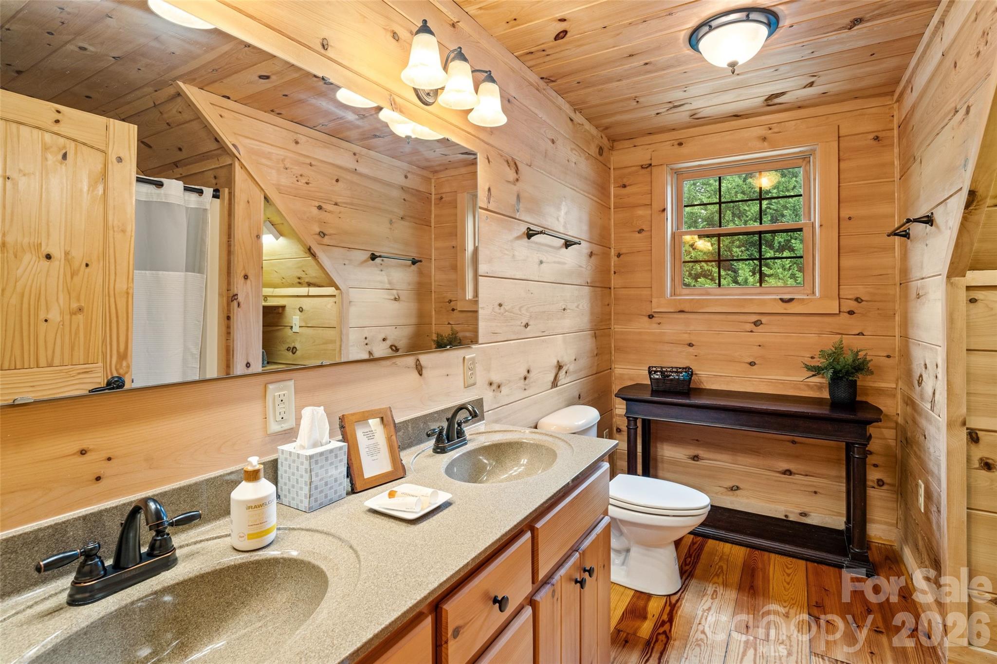 One of two huge upstairs full bathrooms, complete with a dual-sink vanity, ample counter space, full-width mirror with multi-bulb vanity lighting, character wood finishes, and a window framing wooded views. - 213 Big Valley Road, Pisgah Forest