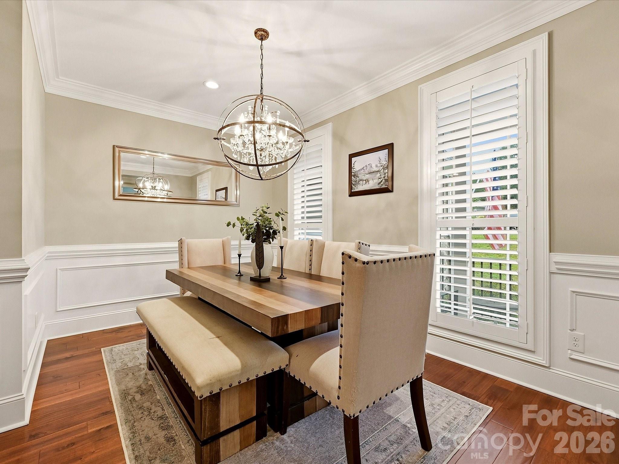 An elegant formal dining room features rich floors, classic wainscoting and crown molding, plantation shutters, and a striking statement chandelier. - 3167 James Plantation Drive, Denver