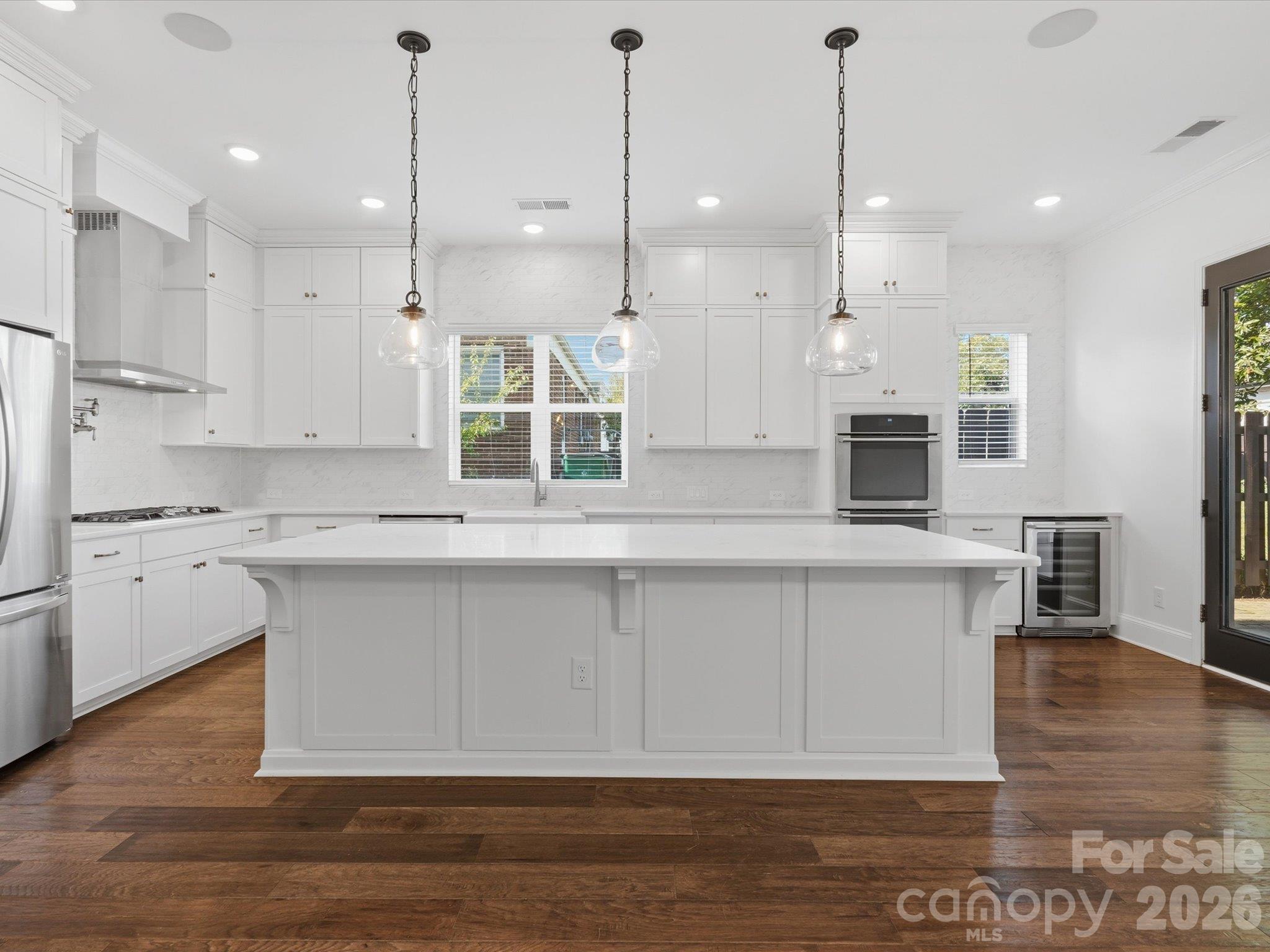White chef's kitchen with oversized entertaining island, glass pendant lights, stainless French-door refrigerator, stainless chimney range hood, double wall ovens, and wine refrigerator. - 337 Belton Street, Charlotte