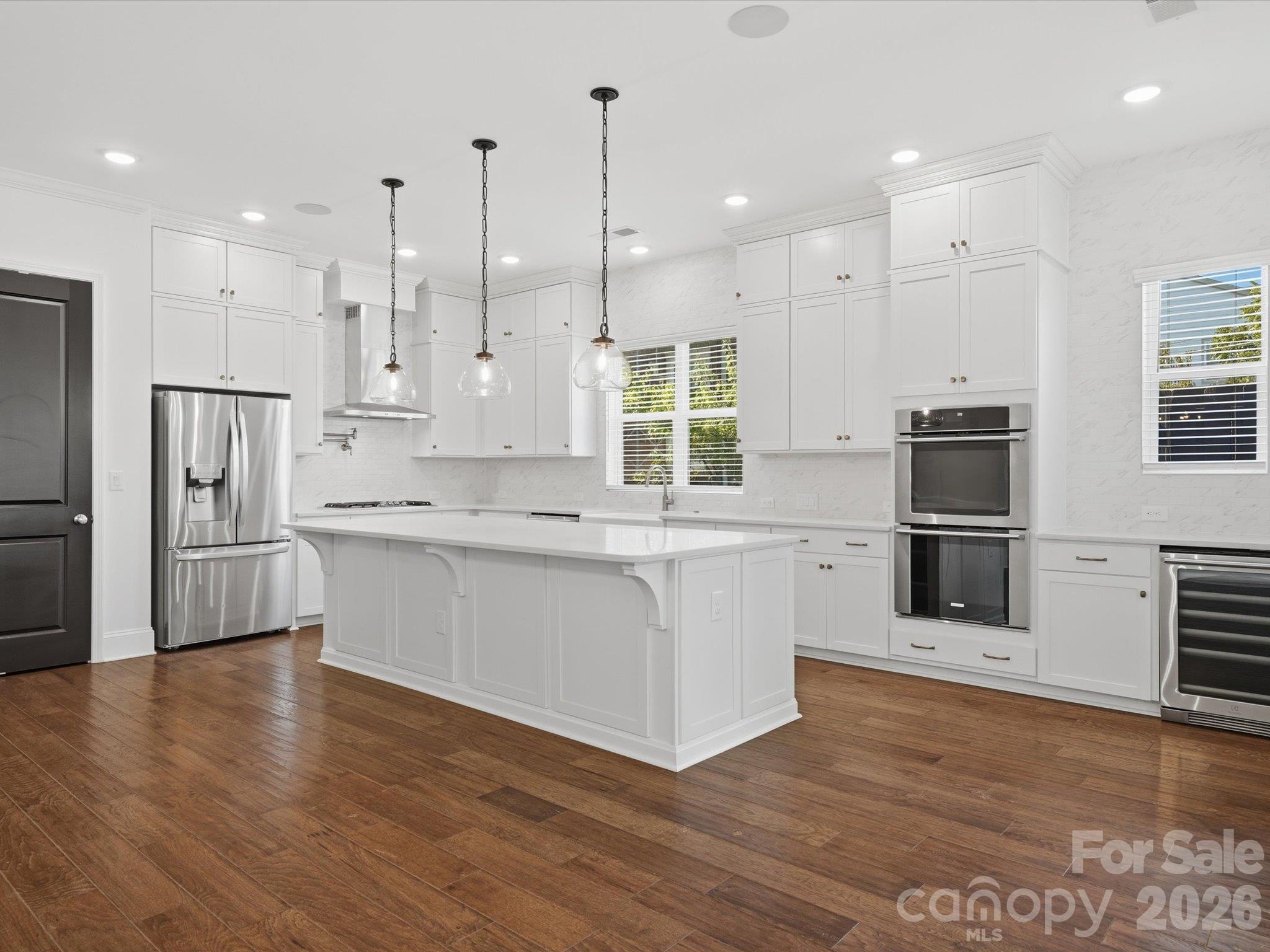 Kitchen with oversized island, floor-to-ceiling white shaker cabinets, gas cooktop with stainless chimney hood, built-in double ovens, under-counter wine fridge, and recessed lighting. - 337 Belton Street, Charlotte