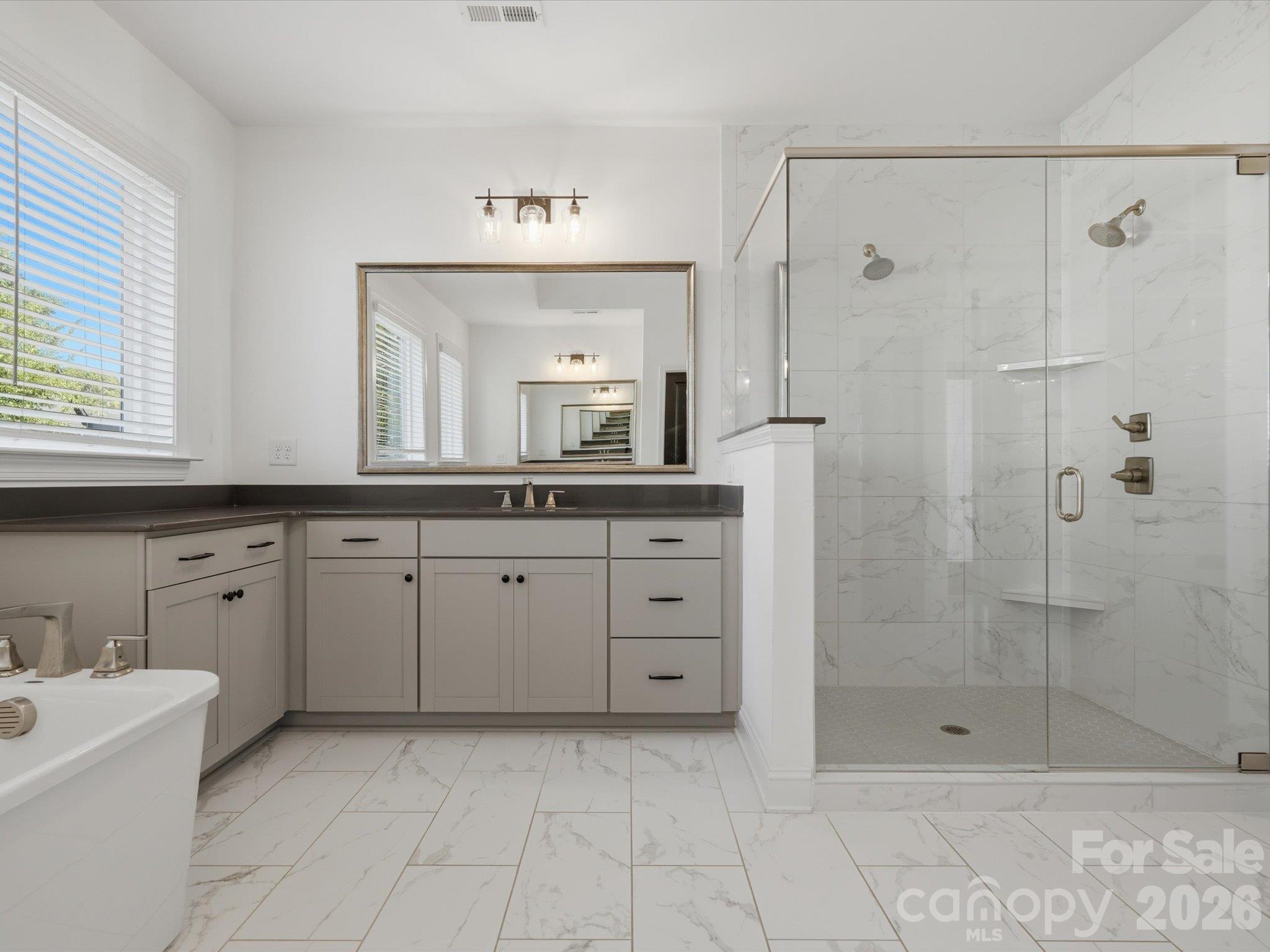 Primary bath with frameless glass walk-in shower, marble-look tile surround, dual showerheads, and gray shaker vanity with dark countertop. - 337 Belton Street, Charlotte