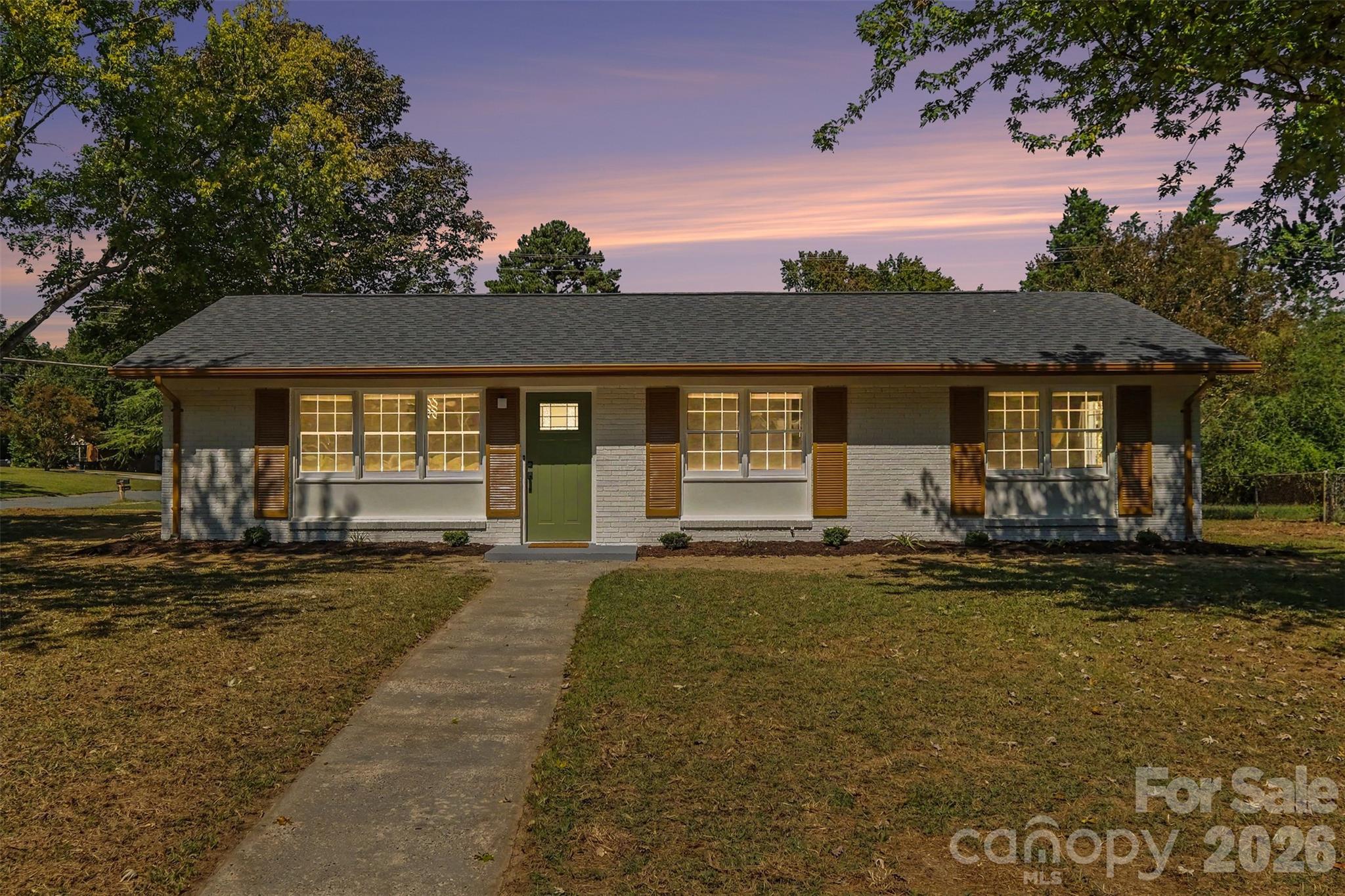 Charming ranch-style home at twilight, showcasing a newly painted brick and siding exterior, brand-new roof and gutters, a welcoming new front door, and manicured landscaping under a vibrant sunset sky - 3801 Hillock Court, Charlotte
