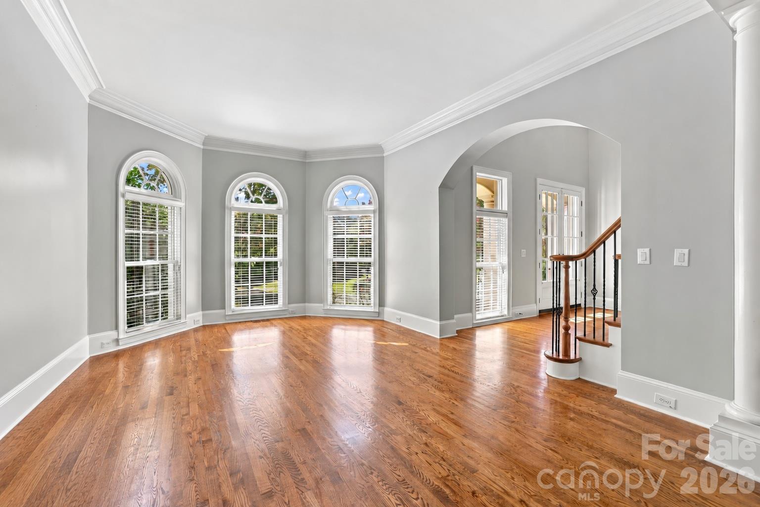 A large bay window with three arched windows brings plenty of natural light into this spacious living room, with hardwood floors and crown molding continuing the home’s classic architectural style. - 2309 Highland Forest Drive, Waxhaw