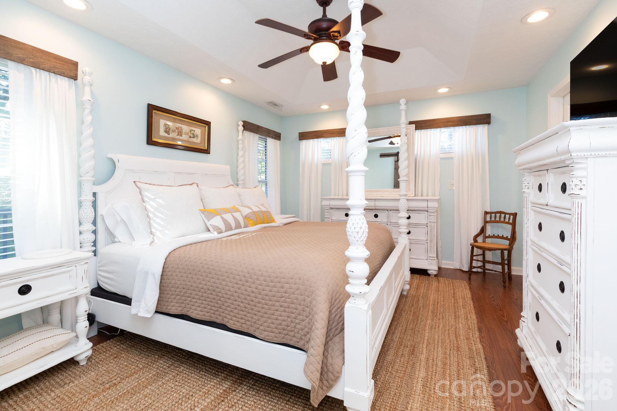 Spacious primary bedroom with a tray ceiling, recessed lighting, ceiling fan, wood flooring, and multiple windows bringing in natural light. - 139 Lone Eagle Lane, Hendersonville