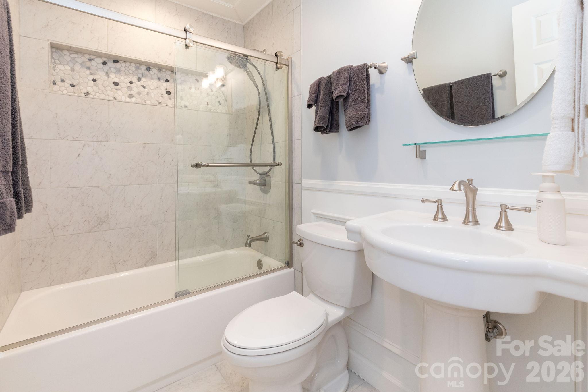 Full bathroom with a pedestal sink, round mirror, and a tub/shower combination accented by tile surround and sliding glass doors. - 139 Lone Eagle Lane, Hendersonville