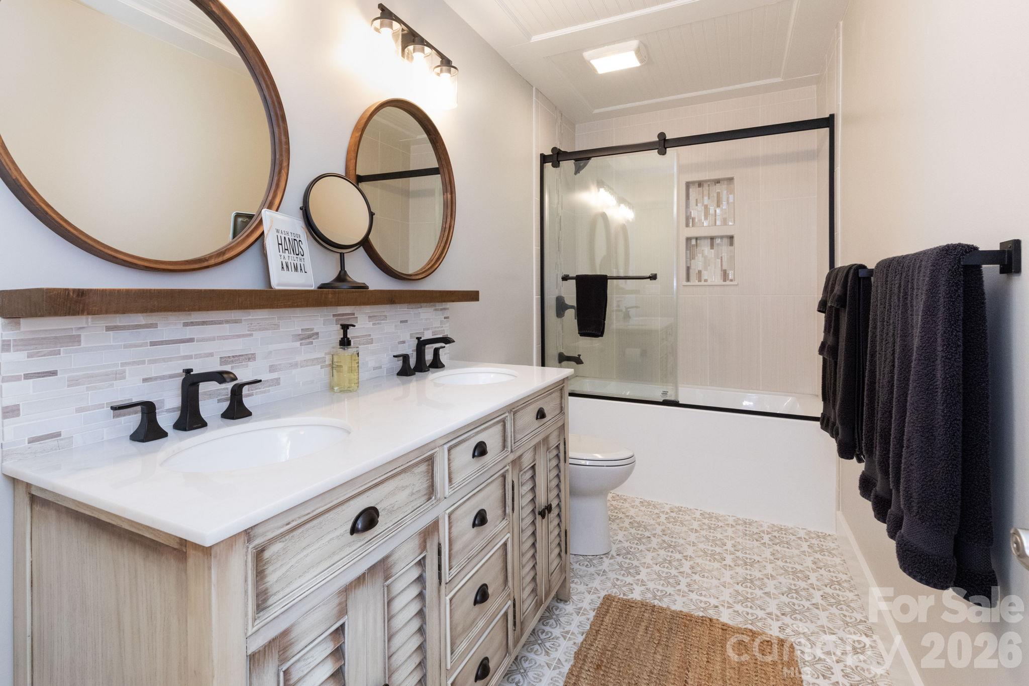 Basement bathroom featuring a double-sink vanity, round mirrors, decorative tile backsplash, and a tub/shower combination with sliding glass doors. - 139 Lone Eagle Lane, Hendersonville