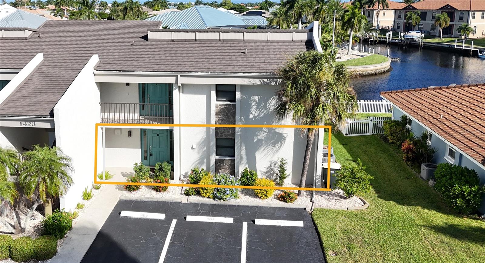 Front aerial of the two-story condo building in Punta Gorda, FL, with the first-floor unit highlighted, paved parking in front, and canal views beyond. - 1423 MEDITERRANEAN DR #B, PUNTA GORDA