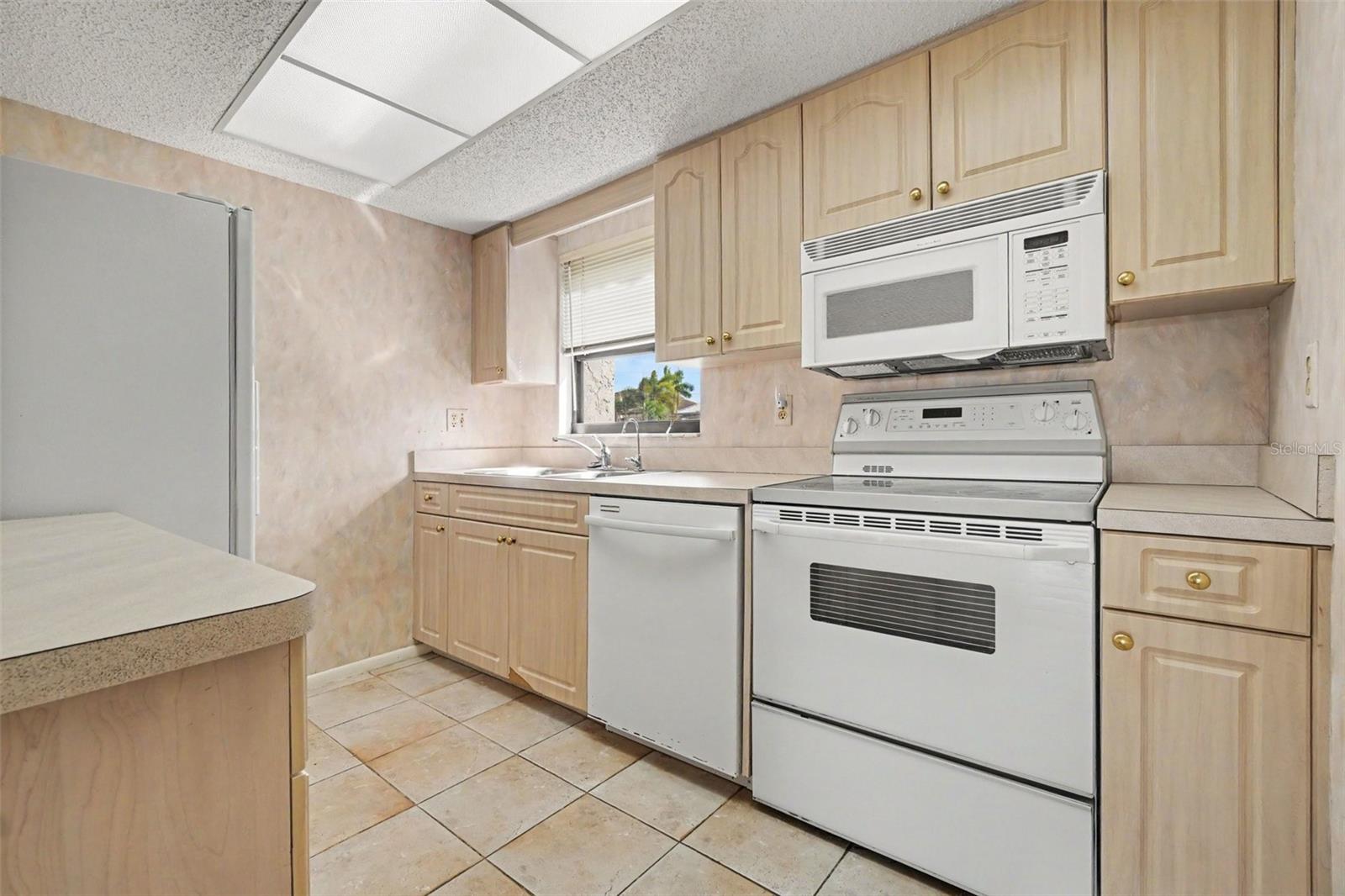 Kitchen with light wood cabinetry, white appliances including a range and over-the-range microwave, tile flooring, and a window over the sink. - 1423 MEDITERRANEAN DR #B, PUNTA GORDA